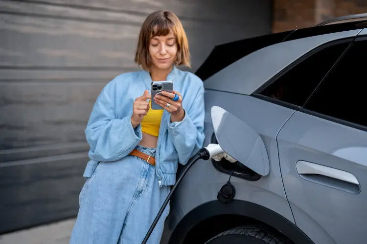 Dekoratives Bild. Eine Frau mit blondem Schulterlangen Haarschnitt schließt ihr parkendes Elektrofahrzeug an einer Elektro-Ladestation an.