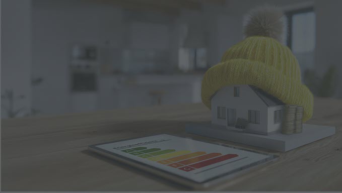 Auf einem Holztisch steht ein Modelhaus, das eine gelbe Pudelmütze trägt. Daneben ein Energielabel. Im Hintergrund eine modern eingerichtete Küche.